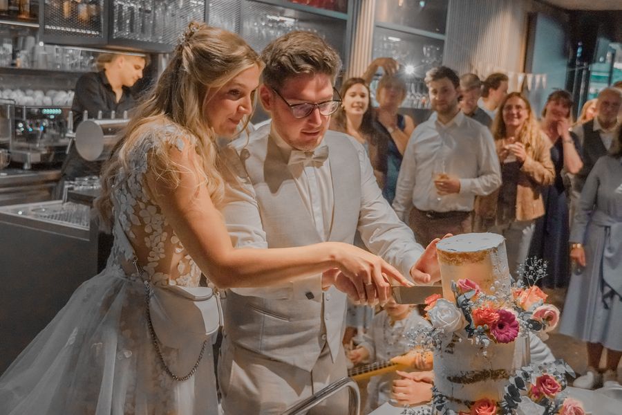 Brautpaar schneidet die Hochzeitstorte an, umringt von applaudierenden Gästen
